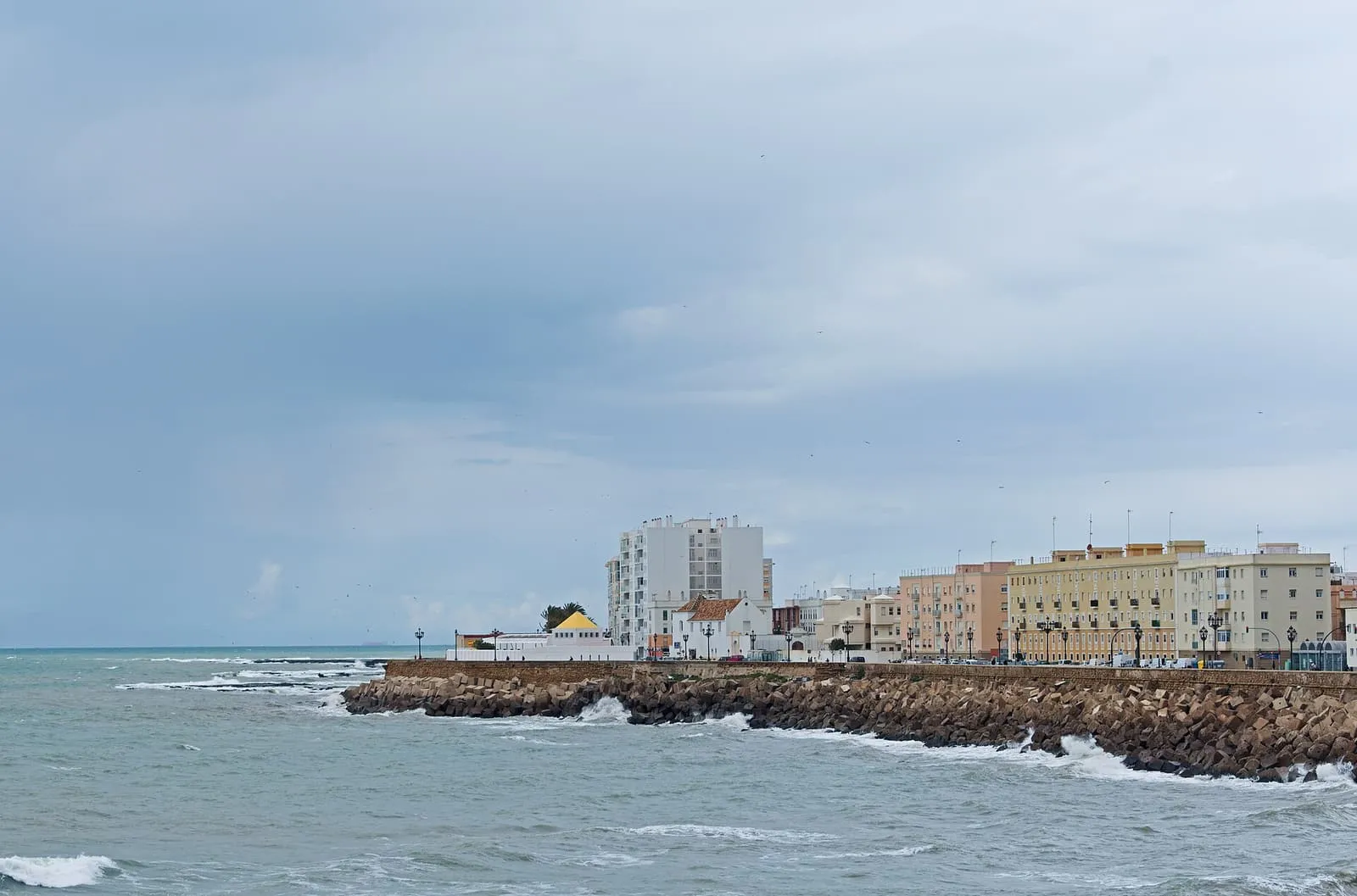 Wellen brechen an der felsigen Uferbefestigung des Campo del Sur in Cádiz, dahinter Häuser am Meer