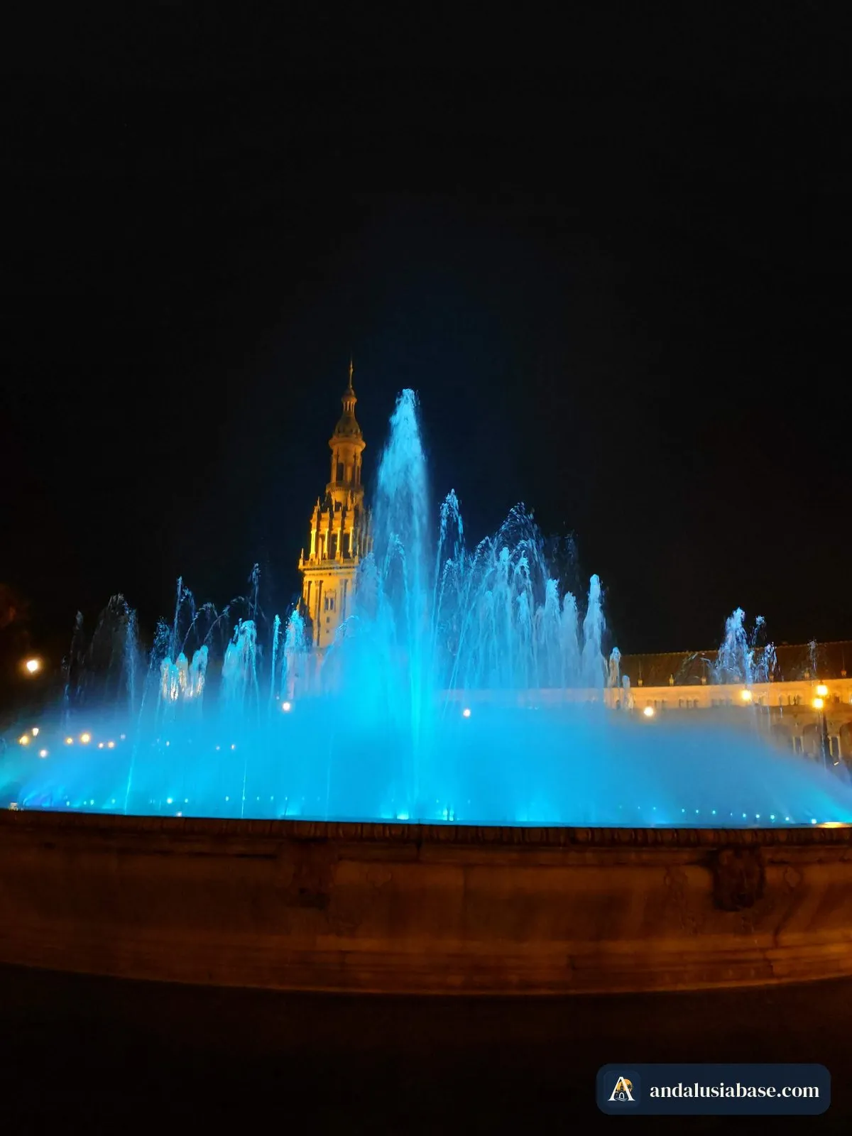 A fonte central iluminada de azul à noite, com uma torre iluminada ao fundo