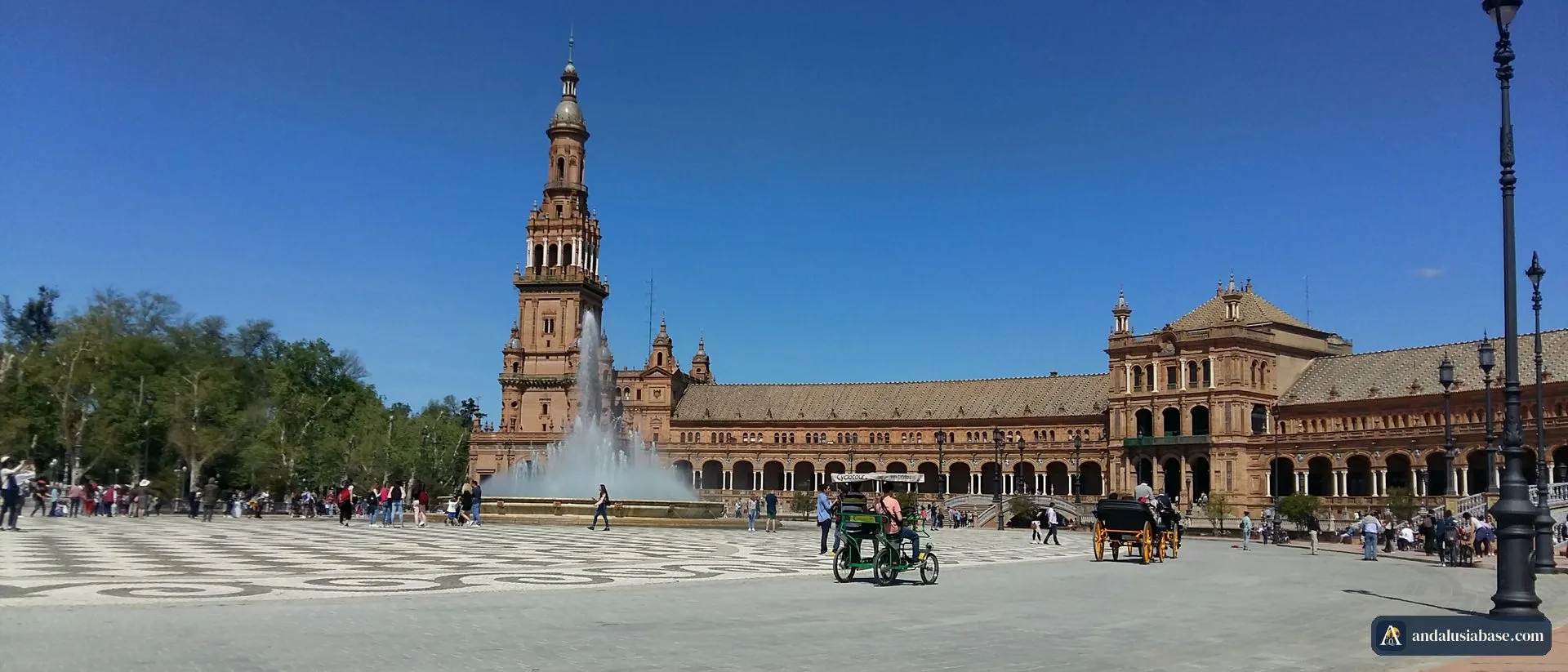 Uma carruagem atravessa a Plaza de España sob o sol de abril, com a torre e a fonte ao fundo