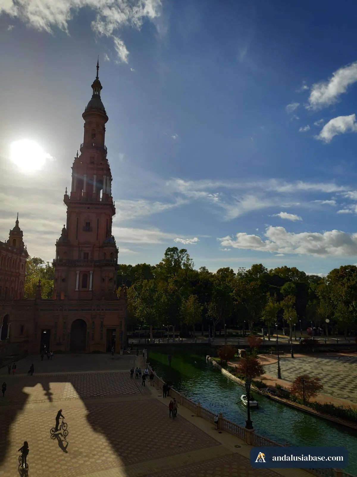 The sun blazing beside a Plaza de España tower, with long shadows across the square