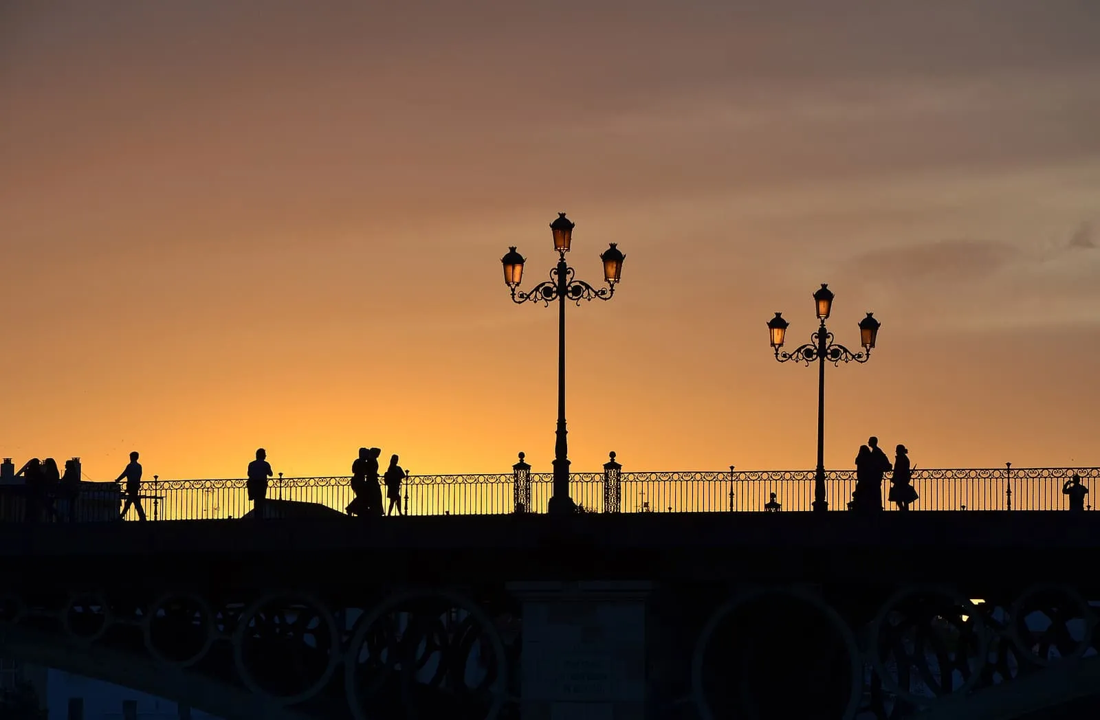 Silhouetten von Passanten und Laternen auf der Triana-Brücke vor orangem Abendhimmel, Sevilla